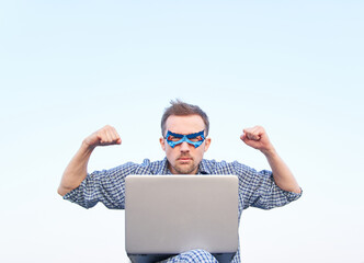 Pumped fists or superpower, serious emotion concept. Caucasian male in nightwear shirt and superhero blue mask using laptop and showing strength gesture. High quality image