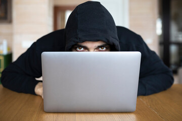 Portrait of enigmatic man in hoodie sits at the desk and works on laptop