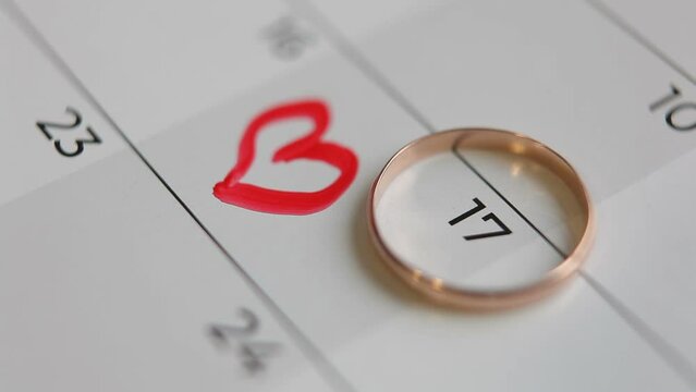  The wedding ring lies on the calendar date of the 17th. A man or woman draws a heart with a red marker on a calendar. Round date of acquaintance or wedding.