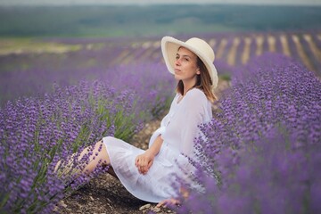 A young relaxed girl breathes fresh air while sitting in a hat in a lavender field on a sunny day.