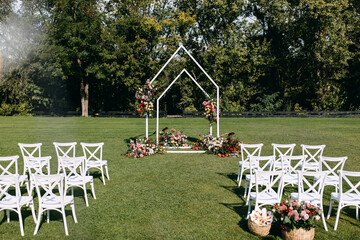 Wedding ceremony aisle with an arch decorated with flowers and white wooden chairs placed on green...