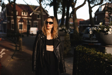 Pensive blonde woman wear black dress, glasses, leather jacket and touch it, posing. Outdoor shot of calm sensual hippie lady with two thin braids and wave hair. Coachella or boho freedom style.