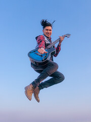 Hombre joven brincando mientras toca su guitarra, cielo azul de fondo 