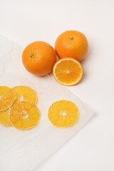 Two fresh oranges, next to which are dried orange slices on parchment paper. White background.