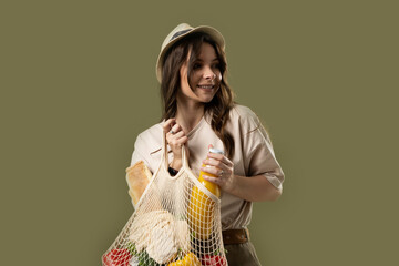 Smiling young woman in light summer clothes with a mesh eco bag full of vegetables, greens watching in a camera on a green studio background. Sustainable lifestyle. Eco friendly concept. Zero waste.