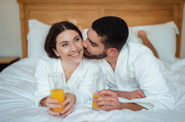 Obraz na płótnie Canvas Boyfriend Kissing Girlfriend Holding Juice Lying Relaxing In Bed Indoors