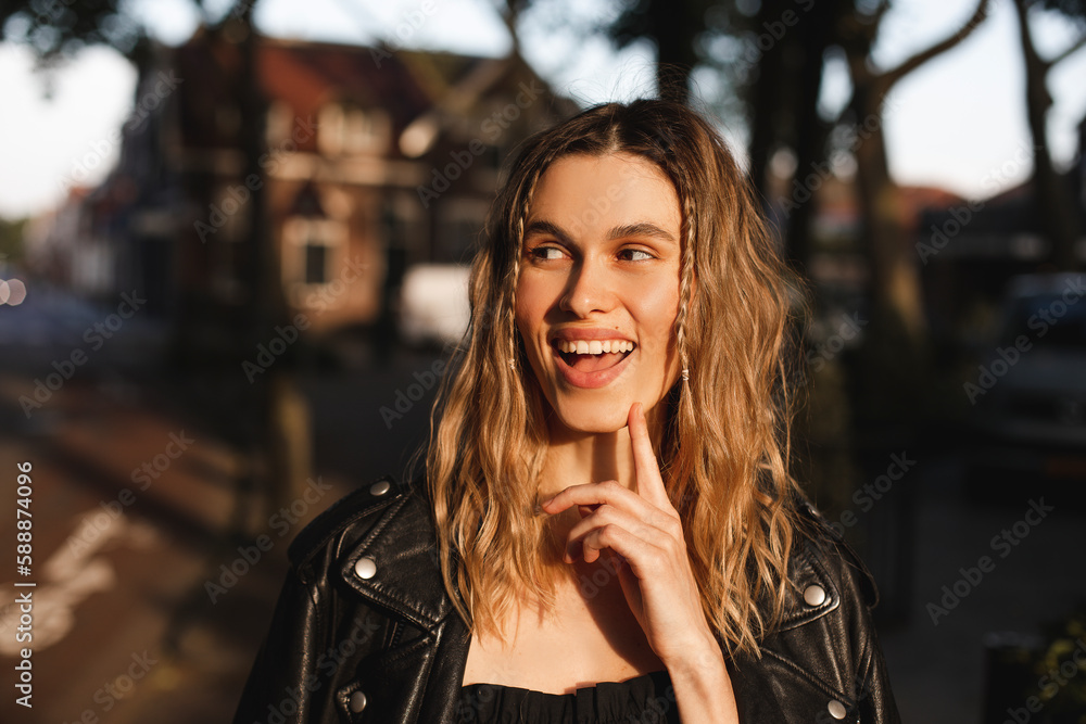 Poster Pensive blonde woman in black leather jacket and dress with open mouth and finger near face, good idea. Outdoor shot of happy hippie lady with two thin braids and wave hair. Coachella freedom style