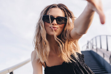 Cheerful lady making selfie at sunny day. Pretty young girl with curly blonde hair and two thin braids towards to camera, smiling and wear black glasses and dress, send blow kiss under sky and stairs.