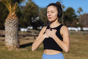 Concentrated young woman wearing headphones doing yoga meditation with online tutorial outdoors at...
