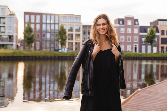 Pensive blonde woman wear black dress, leather jacket and touch it, posing, turn around. Outdoor shot of happy hippie lady with two thin braids and wave hair. Coachella or boho freedom style.