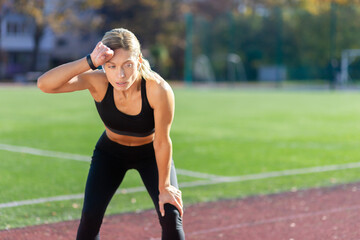 Tired sportswoman resting after jogging on a sunny day at the stadium, blonde in sportswear taking...