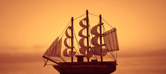 Miniature ship model on orange sunset sky, yellow sea. sailboat model. Pirate ship sailing on the ocean at sunset. - Powered by Adobe