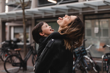 Fototapeta na wymiar Happy young woman have fun with cute child boy holding him on arms and hug. Mommy little kid son together outdoors. Mother's Day love family. Woman with boy go to home. Mom and son show tongue, play.