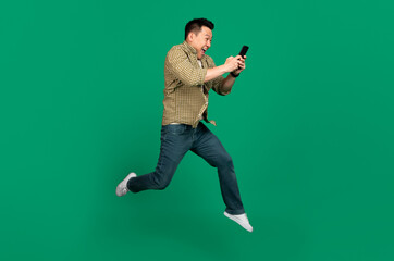 Full length of asian man with smartphone jumping up over green studio background, copy space