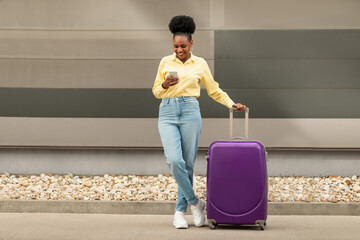 Cheerful Black Woman With Travel Suitcase Using Phone Texting Outdoors