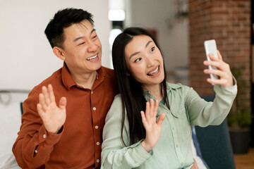 Fototapeta na wymiar Portrait of cheerful asian married spouses using cellphone video calling and waving hello to phone webcamera