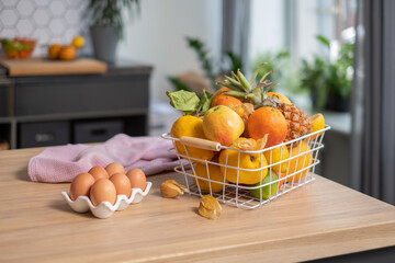 Juicy fruits in a metal basket, eggs in a stylish white dish and a linen napkin on a light wooden table. A shelf and a green houseplant are in the blurred background.