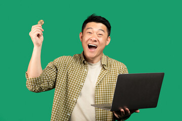 Overjoyed asian businessman holding laptop and bitcoin coin, posing, looking at camera with excitement, green background