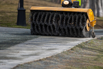 A mechanical rotating street cleaning brush attachment for a truck on a paved sidewalk cleans the dirt and debris with its large metal coils. The metal industrial sweeper is yellow in color.
