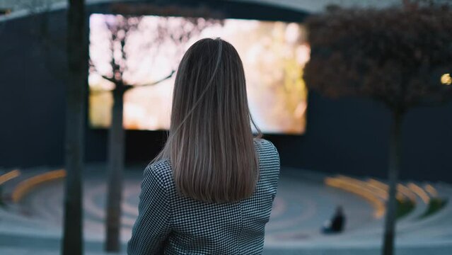 Rear view of a young woman watching a movie or broadcast on a big screen in a street cinema