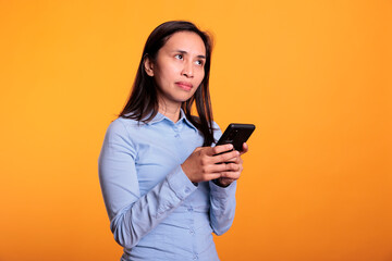 Filipino woman chatting with remote friend, typing message on mobile phone in studio over yellow background. Cheerful attractive young adult browsing information on internet working at project