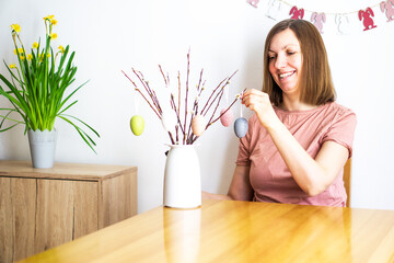 Smiling woman decorating a bouquet of willow branches with colored Easter eggs at home. Happy Easter
