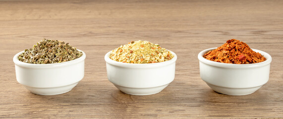 Three white saucers with various bright spices stand in a row on a wooden table.