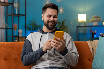 Caucasian man sitting on couch uses mobile phone smiles at home living room apartment. Young guy texting share messages content on smartphone social media applications online, watching relax movie