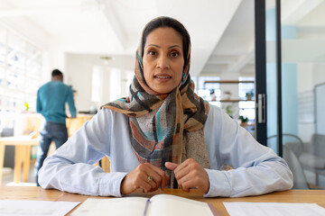 Portrait of happy muslim biracial businesswoman in headscarf looking at camera in modern office
