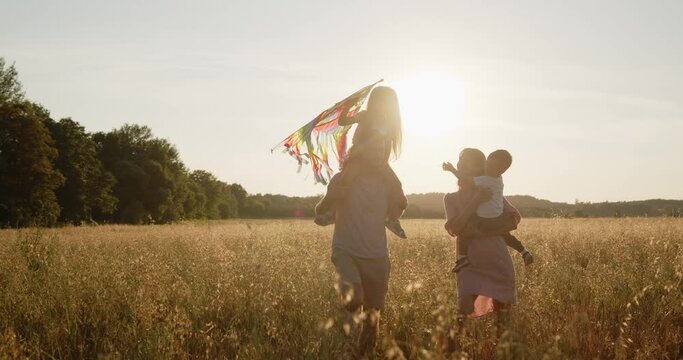Happy young smiling family of four have fun in a golden wheat field at sunset. Mom, dad, son and daughter walk together holding colorful kite during golden hour.