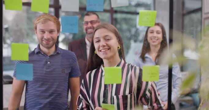 Work colleagues celebrating successful startup project. Caucasian business team at glass wall with sticky notes. Scrum with colorful stickers. Yeah gestures with hands up.