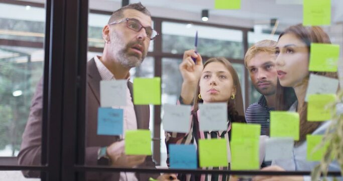 Work colleagues discussing startup project ideas. Team of caucasian business people standing at glass wall with sticky notes. Scrum methodology with colorful stickers. Left camera movement.