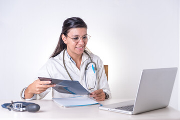 A female doctor in a white coat is sitting at a desk with an X-ray in her hand talking on the laptop.