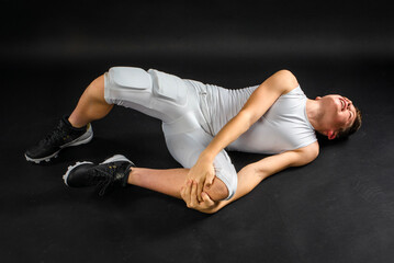 Young preteen football boy in football uniform laying injured on studio floor with black background