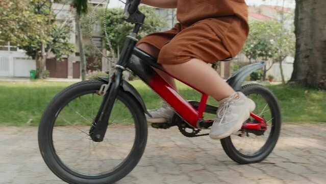 Tilt up shot of little Asian kid in protective helmet riding bike along city street on summer day
