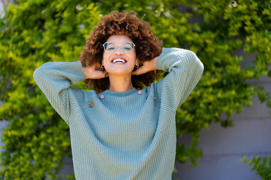 Portrait of biracial cheerful young woman with hands in afro hair smiling and standing in yard