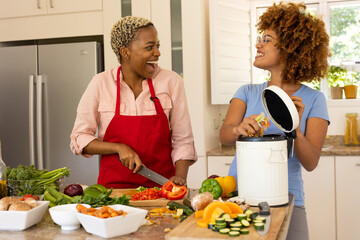Cheerful multiracial lesbian couple chopping vegetables and throwing waste in compost bin in kitchen
