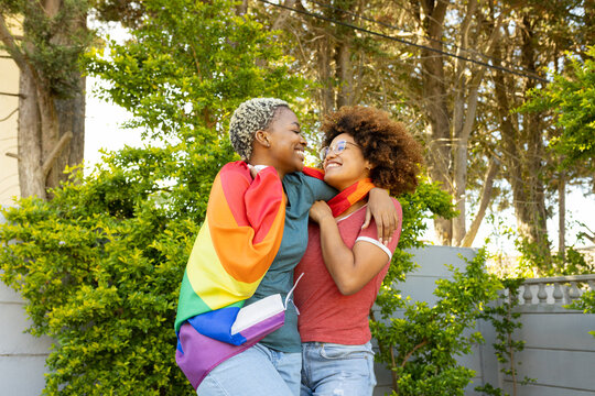 Happy multiracial lesbian couple with arms around embracing rainbow flag against trees in yard