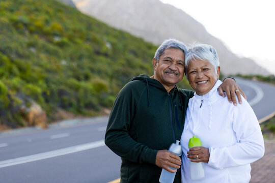 Portrait of happy senior biracial couple wearing sportswear, embracing on street in mountains