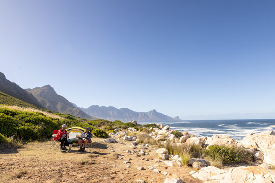 Happy senior biracial couple sitting on camping chairs and drinking coffee, with copy space