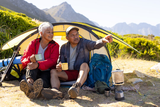 Happy senior biracial couple sitting in tent and drinking coffee in mountains