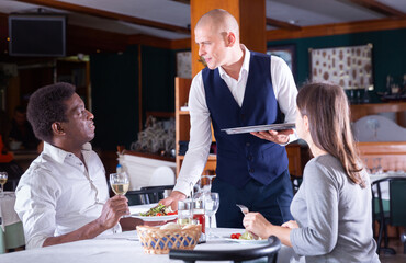 Polite waiter serving adult biracial couple in restaurant, bringing ordered meals