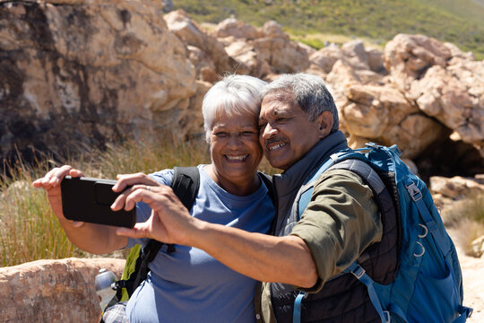 Happy senior biracial couple wearing backpacks, hiking and taking selfie
