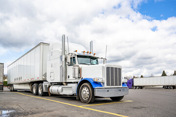 Stylish white big rig semi truck with blue accents and loaded dry van semi trailer standing on the market spot on truck stop parking lot for truck driver rest