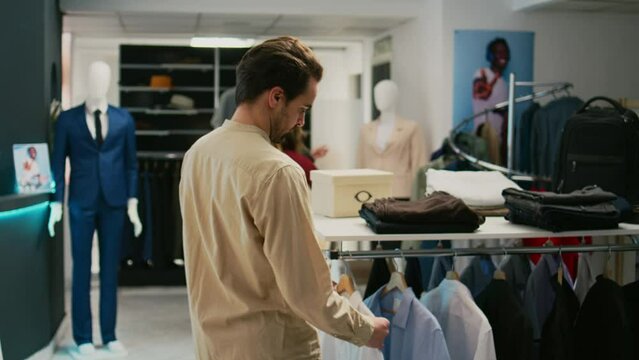 Modern man shopping for formal wear in clothing store, young adult looking at hangers filled with trendy clothes. Retail shop customer examining boutique merchandise on display, small business.