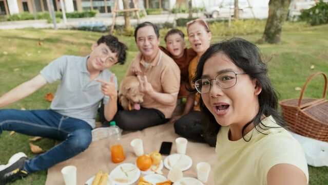 POV of happy Asian woman holding camera and posing for selfie with family and cute dog on summer picnic in park
