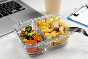 Container with tasty food, fork and laptop on white wooden table, closeup. Business lunch