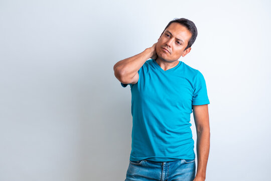 Hombe latino tocandose el cuello muy serio con playera y pantalon de mezclilla azul y fondo blanco