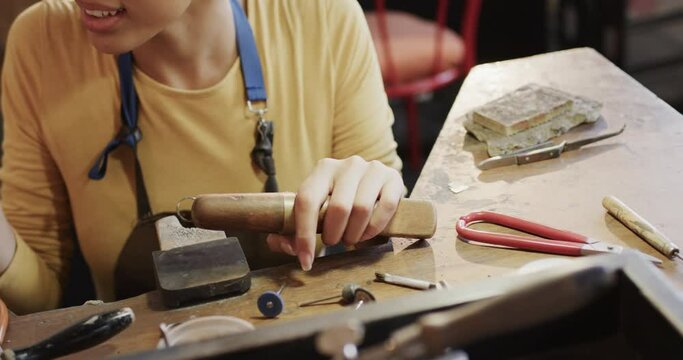 Happy biracial female worker at table shaping ring with handcraft tools in studio in slow motion
