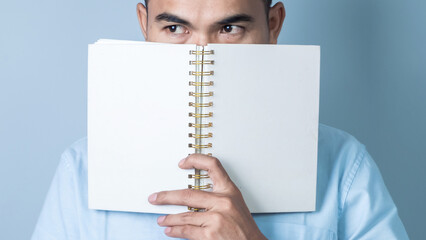 Confident Asian man covering his face with notebook on light blue background